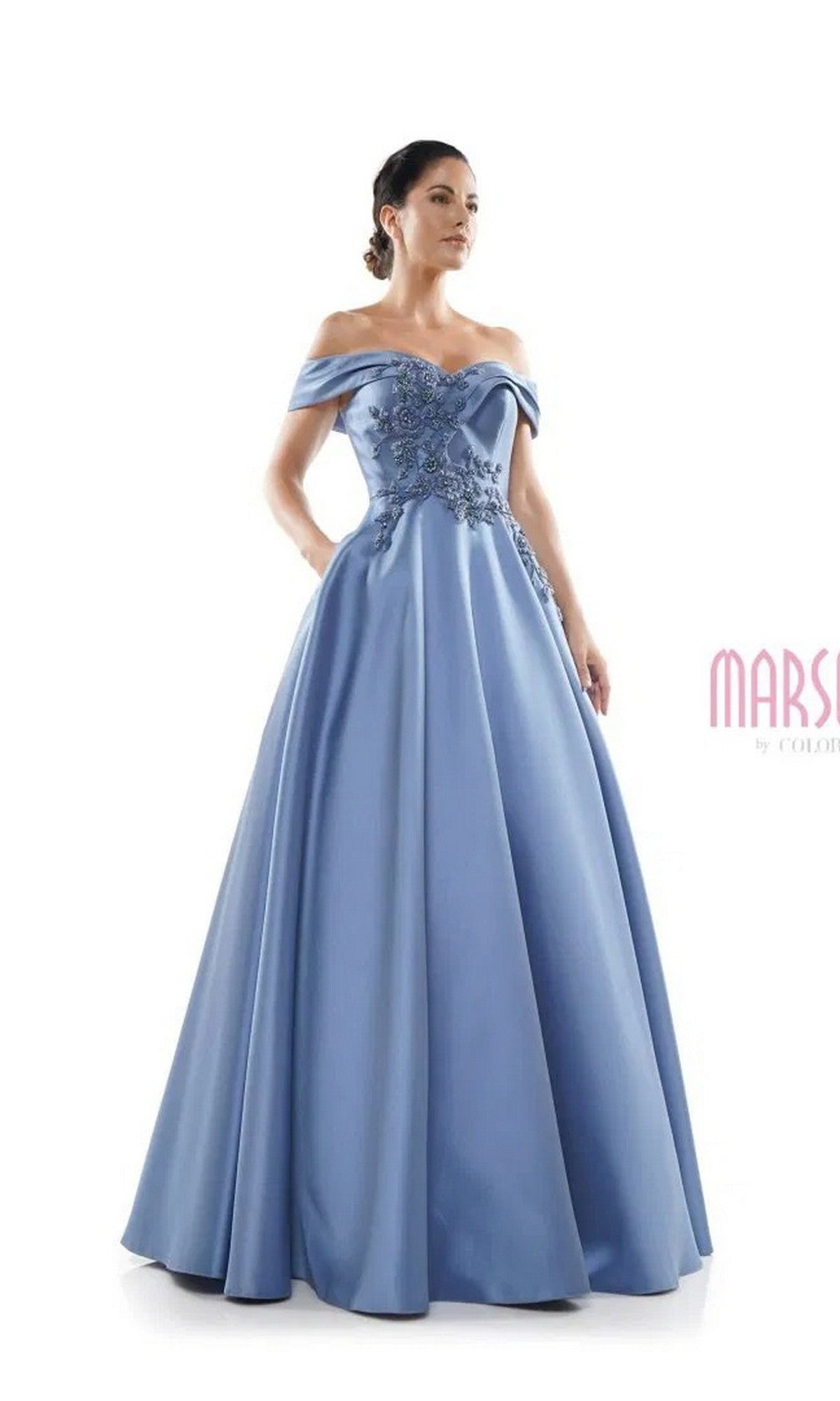 A woman stands against a plain white background wearing the Marsoni MV1008, a long formal off-the-shoulder light blue gown with floral embroidery and a fitted bodice.